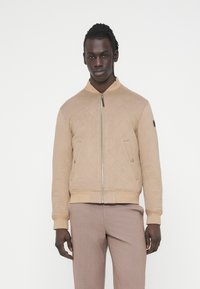 Μπεζ μπουφάν bomber με κουκούλα, φερμουάρ κλεισίματος και πλαϊνές τσέπες. Πλεκτές μανσέτες και τελείωμα, διακοσμημένα με ένα έμβλημα λογότυπου.