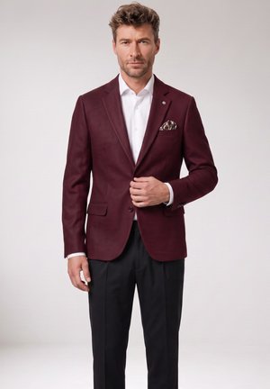 Man wearing a burgundy blazer with patterned pocket square, white shirt, and black pants, standing against a plain light background.