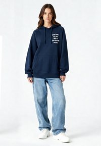 Sweat à capuche bleu marine avec texte blanc, poche kangourou à l'avant et capuche avec cordon, associé à un jean ample bleu clair et des baskets blanches.