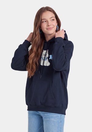 Giovane donna con capelli lunghi che indossa orecchini a cerchio blu, una felpa blu navy con toppa di orso bianca e jeans azzurri, che guarda leggermente di lato.