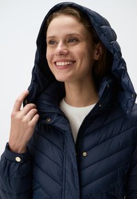 Giacca puffer blu navy con cappuccio, dotata di chiusura con zip, accenti in hardware dorato e motivo a rombi trapuntato. Texture morbida.