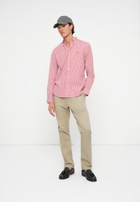 Man wearing gray cap, red and white striped button-up shirt, beige pants, and black tassel loafers, standing against a white background.
