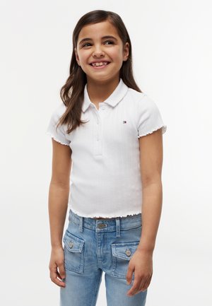 Smilende pige med langt brunt hår iført en hvid kortærmet polo-shirt og blå jeans med lommeklapper, stående mod en hvid baggrund.