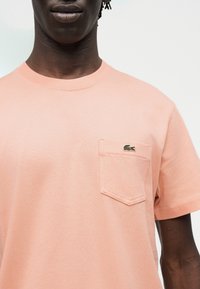 Persona que lleva una camiseta de manga corta color rosa claro con un bolsillo en el pecho que muestra un pequeño logo de cocodrilo verde.