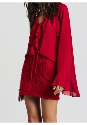 Vrouw draagt een rode blouse met ruches aan de voorkant en uitlopende mouwen, gecombineerd met een gestructureerde rode minirok met bloemmotieven.