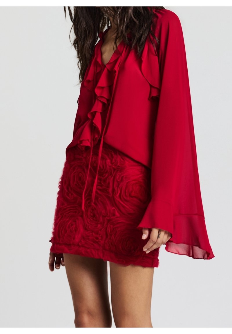 Femme portant un chemisier rouge à volants sur le devant et manches évasées, associé à une mini-jupe rouge texturée à motifs floraux.