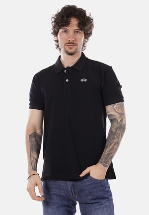 Uomo con capelli ricci che indossa una polo nera e jeans blu, mostra tatuaggi dettagliati su entrambe le braccia, in piedi con una mano in tasca.