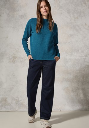 Pull en maille texturée de couleur sarcelle avec des manches longues, associé à un jean sombre décontracté et des baskets de couleur claire. Fond uni.