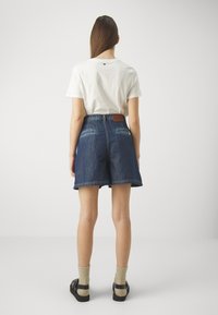 Camiseta blanca de manga corta, pantalones cortos de mezclilla con un parche de cuero marrón en la cintura y sandalias negras con calcetines grises. Diseño sencillo, ajuste casual.