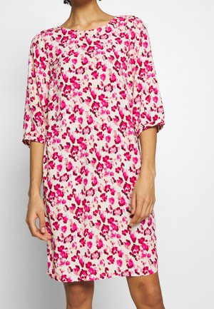 Femme portant une robe ample, longueur genoux, avec des motifs floraux roses et rouges et des manches trois quarts sur fond blanc.