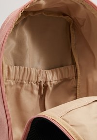 Interno di uno zaino in tessuto rosa con rivestimento beige, tasca elastica e texture liscia. Presenta una tasca laterale in rete con chiusura a zip.