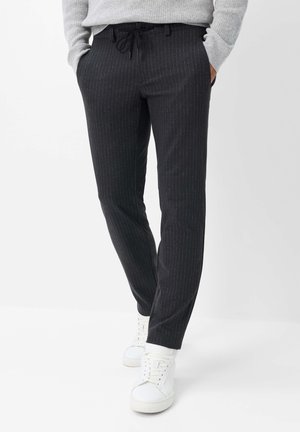 Person trägt schwarze Nadelstreifenhose, hellgrauen Pullover und weiße Turnschuhe, steht mit Händen in den Taschen vor weißem Hintergrund.