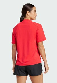 Röd kortärmad skjorta i bomull med rund halsringning, prydd med en logotyp vid fållen och en slät yta. Bärs med svarta shorts.