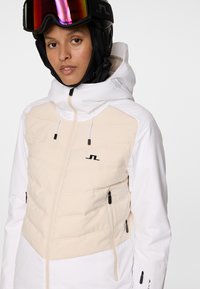 Młoda osoba w beżowej i białej kurtce narciarskiej z kapturem oraz czarnym kasku narciarskim z lustrzanymi goglami na tle białym.