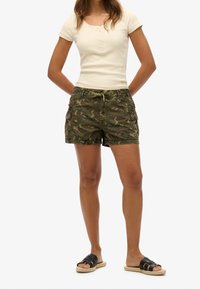 Pantalones cortos cargo de camuflaje con múltiples bolsillos, de longitud hasta la mitad del muslo y cintura ajustable con cordón. Combinados con una camiseta beige clara de canalé y sandalias.