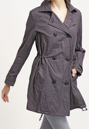 Femme portant un trench-coat gris foncé à double boutonnage avec des boutons, des poches latérales et des détails ajustables à la taille et aux manches.