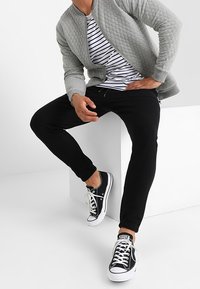 Giacca bomber trapuntata grigia sopra una t-shirt a righe bianca e nera, abbinata a pantaloni slim neri e sneakers basse nere con suole bianche.