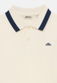 Kremt polo-skjorte med en marineblå ribbestrikket krage, med tre knapper og en liten marineblå logo på venstre side. Strukturert stoff.