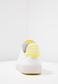 Vit sneaker med en texturerad meshovandel, gul hältab och gummisula med ett mönstrat design. "Pharrell Williams"-logotyp på baksidan.