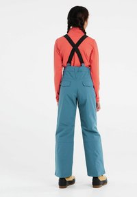 Blå vattentäta overaller med hög midja och justerbara svarta hängslen, tillsammans med en långärmad koralltop och beige stövlar.