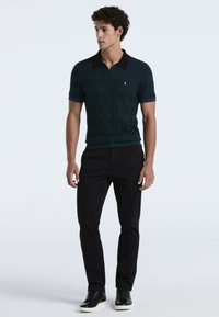 Jonge man staat in een donkerblauw gemarmerd poloshirt met korte mouwen en een zwarte kraag, zwarte broeken en zwarte sneakers met witte zolen.