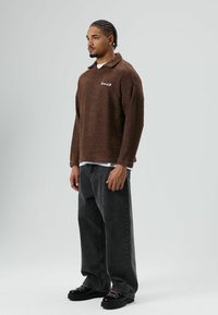 Polaire marron avec col et détail brodé, associée à un jean noir ample et des chaussures noires épaisses. Surface lisse et texturée.