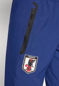 Μπλε αθλητικό παντελόνι με μαύρη τσέπη με φερμουάρ και πατς με λογότυπο πουλιού και το κείμενο "JAPAN JFA" στο πλάι. Ομαλή υφή.