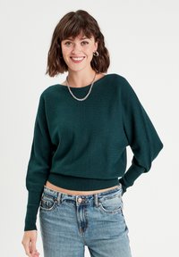 Pull cropped vert foncé avec un large col et des manches longues légèrement bouffantes. Matériel en tricot avec un ourlet côtelé. Porté avec un jean bleu.