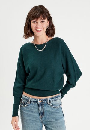 Pullover corto verde scuro con un ampio scollo e maniche lunghe leggermente a sbuffo. Materiale lavorato a maglia con orlo a coste. Indossato con jeans blu.