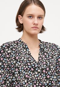 Svart bluse med et blomsterdesign i rosa, blått og hvitt; har V-hals, lange ermer og lett, strukturelt stoff.