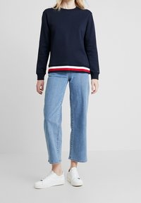 Felpa navy con scollo a girocollo e dettagli a righe rosse e bianche sull'orlo, abbinata a jeans a gamba larga di un blu chiaro e sneakers bianche.