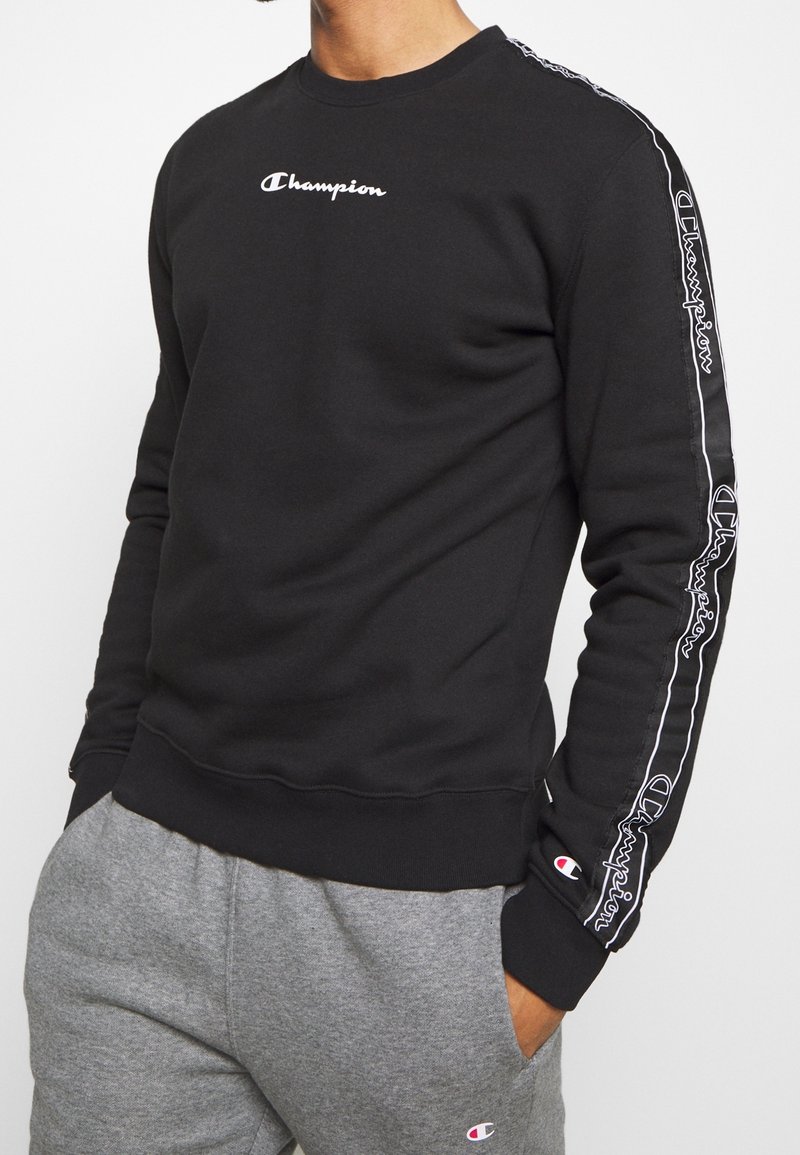Felpa nera con logo "Champion" bianco sul petto. Presenta del nastro laterale bianco con logo ripetuto e polsini a costine. Abbinata a pantaloni della tuta grigi.