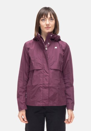 Donna che indossa una giacca impermeabile viola con zip e cappuccio, in piedi contro uno sfondo chiaro uniforme, rivolta verso l'osservatore.