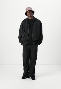 Schwarze Bomberjacke mit Reißverschluss und elastischen Bündchen, kombiniert mit schwarzen weiten Hosen mit Kontrastnähten und einem hellrosanen Bucket Hat.
