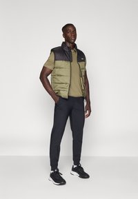 Gilet matelassé en vert et noir avec un col haut, porté sur une chemise ajustée olive ; associé  à un pantalon noir fuselé et des baskets noires.