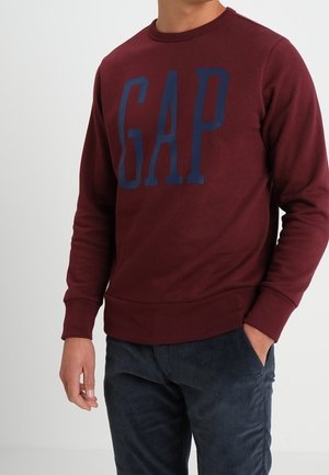 Felpa maroon con logo "GAP" in blu navy. Collo rotondo, maniche lunghe, polsini e fondo a coste, texture morbida, indossata sopra pantaloni in velluto a coste scuri.