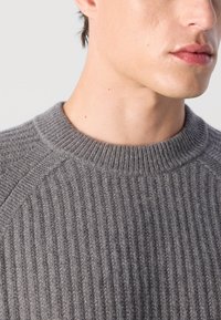 Close-up of a person wearing a gray ribbed knit sweater, focusing on the neck and lower face with a neutral background.