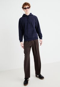 Hanorac fleece de culoare navy cu buzunar tip cangur, purtat împreună cu pantaloni largi maro și adidași negri cu talpă maro. Modelul poartă ochelari de soare negri.