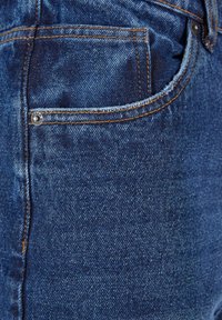 Blå denim jeans med et tæt på billede af stofteksturen, orange syninger og en lille metalnitte på lommen.