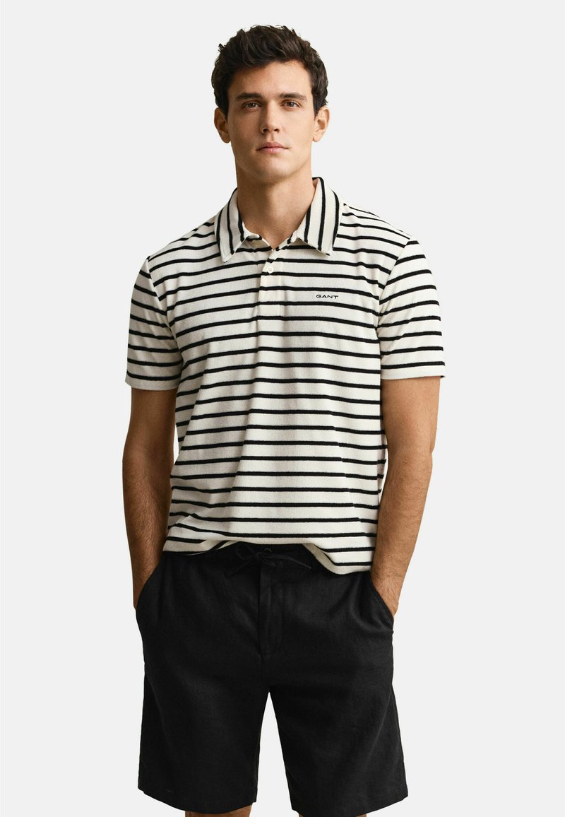 Jeune homme portant un polo à manches courtes à rayures blanches et noires et un short noir, debout les mains dans les poches devant un fond blanc.