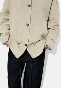 Lys beige jakke med en afslappet pasform, der har brune knapper, to sidelommer og opløftede ærmer, parret med mørke jeans.