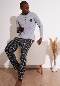 Tricou lung gri cu mâneci lungi, cu butoane negre și logo; pantaloni de pijama cu carouri alb-negru; așezat pe un scaun bej, ținând o cană neagră.