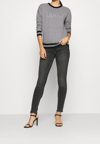 Nainen, jolla on yllään harmaa Liu Jo -neule, mustat kapeat farkut ja mustat korkokengät, seisoo vaihtamatta taustaa vasten.