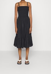 Robe midi noire avec un corsage smocké, de fines bretelles et un ourlet à volants. Associée à des tongs blanches dotées d'une semelle plate-forme noire.