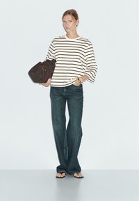 Gestreiftes Langarmoberteil in Creme und Schwarz, kombiniert mit dunklen Jeans. Das Model hält eine braune Tasche und trägt schwarze Flip-Flops. Einfacher Hintergrund.