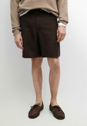 Uomo con pantaloncini marrone scuro, felpa beige, maglietta bianca e mocassini marroni con lacci in piedi davanti a uno sfondo chiaro e semplice.