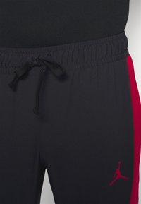 Pantalones deportivos negros con cintura elástica y cordón, que presentan paneles laterales rojos y un logo de Jordan rojo en la parte inferior de la pierna izquierda.