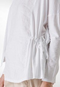 Top bianco a maniche lunghe con vestibilità ampia, raccolto in vita con un cordoncino. Realizzato in tessuto leggero, presenta una finitura testurizzata.