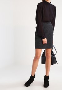 Frau in einer schwarzen Bluse mit langen Ärmeln, grauem knielangen Rock und schwarzen Stiefeletten, die eine schwarze Handtasche an ihrer Seite hält.