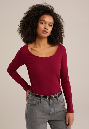 Burgunderfarbener, gerippter Langarmtop mit weiter Rundhals-Ausschnitt, kombiniert mit hochgeschnittenen grauen Jeans, die einen schwarzen Gürtel mit goldener Schnalle tragen.